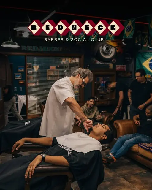 Barbeiro fazendo a barba de um cliente na navalha, sob o letreiro de losangos da Redneck